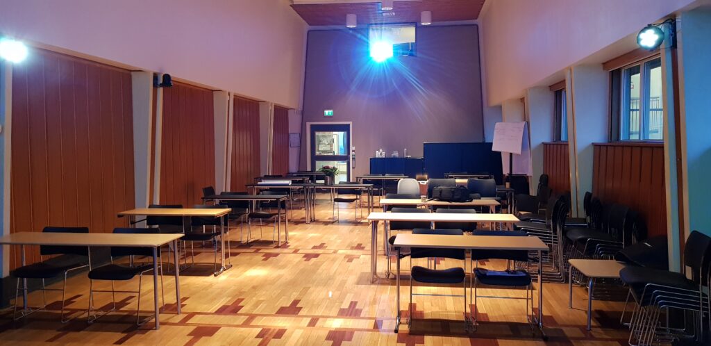 Forsamlingssalen, seen from the lecture podium. One PTZ camera is placed on the left wall and one in the back next to the projector. LED lights help illuminate the speakers.